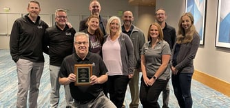 A man holding an award kneels in front of nine adults.