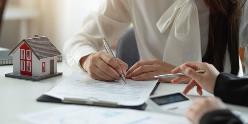 A woman signs a document while another person's hand points to a line on the paper. 