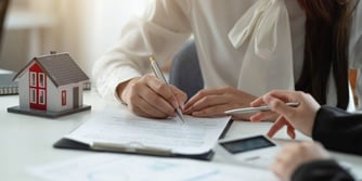 A woman signs a document while another person's hand points to a line on the paper. 