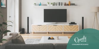 A tv is atop a tv stand behind a coffee table and underneath a bookshelf. 