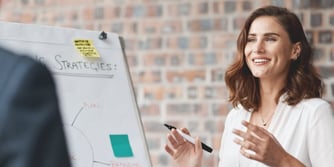 A woman smiles while holding a pen in front of a board outlining strategies in an office. 