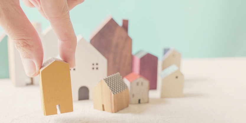 A hand picks up a small, yellow, wooden house from a table with eight small wooden houses.