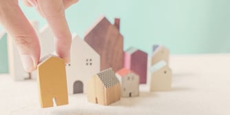 A hand picks up a small, yellow, wooden house from a table with eight small wooden houses.