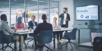 A man stands in front of a visual presentation at the head of a conference table talking to four individuals.