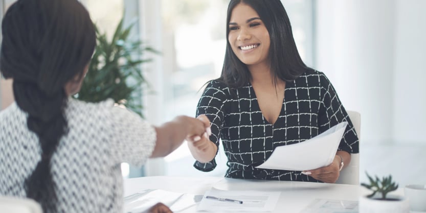 A smiling woman shakes hands with another woman while holding a piece of paper in her other hand.