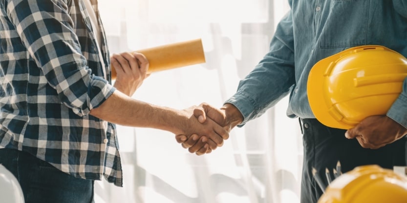Two people shake hands while holding hard hats