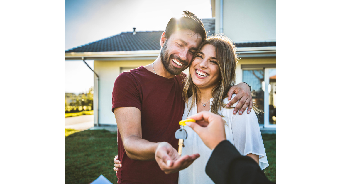 A smiling couple standing in front of a house is handed keys.